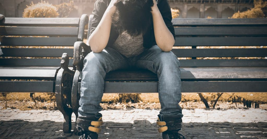 Man in Black Shirt and Gray Denim Pants Sitting on Gray Padded Bench embracing sadness